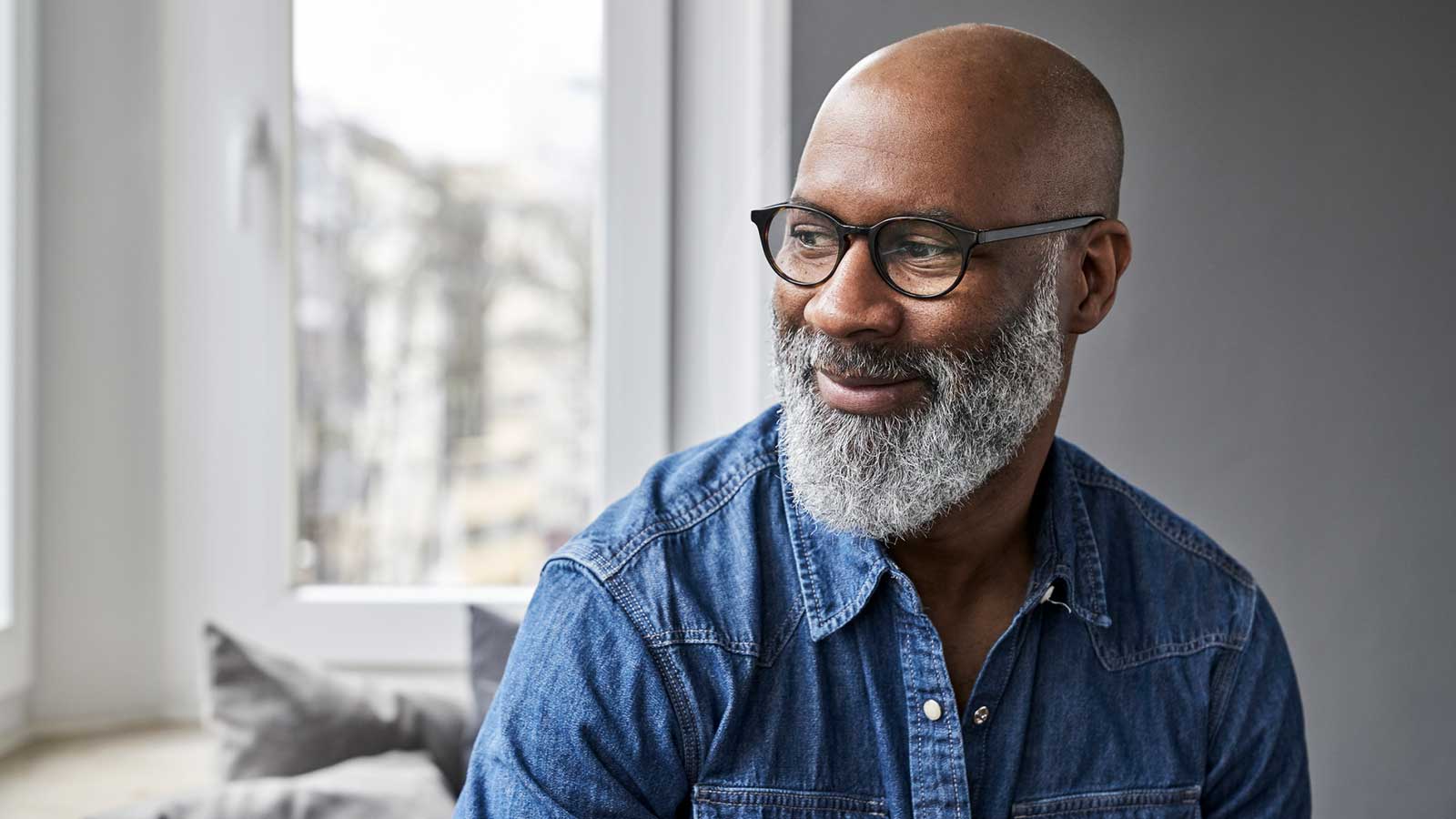 A man with a gray beard and glasses smiles while looking to the side, wearing a denim shirt, sitting by a window with a blurred city view.