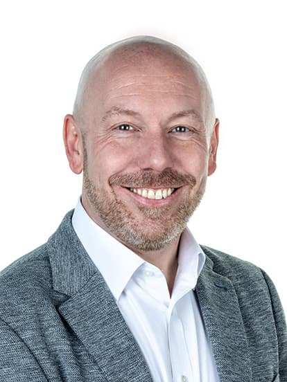 Smiling bald man with a beard, wearing a gray suit jacket and white shirt