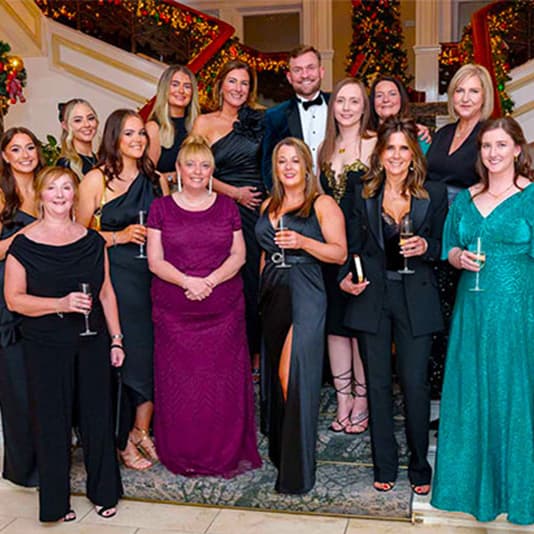 A group of people dressed in formal attire, posing on a staircase decorated with Christmas lights and garlands, holding drinks.