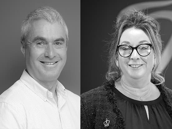 A grayscale image of a man and a woman smiling. The man wears a striped shirt, and the woman wears glasses and a dark top.