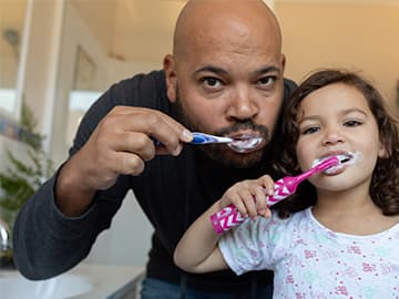 Father and daughter brushing teeth together at bathroom mirror
