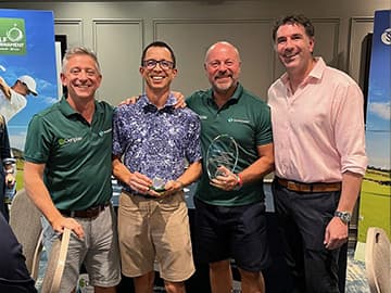 Four men smiling, two holding glass awards, standing indoors. Three wear green shirts, one in a floral shirt, and one in a pink shirt.
