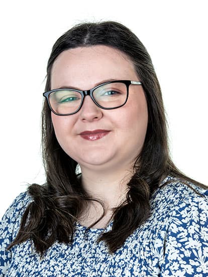 A woman with glasses and long dark hair smiles at the camera, wearing a blue floral-patterned top
