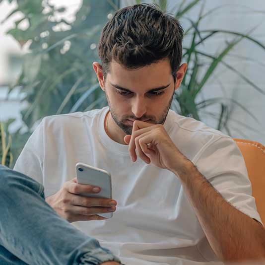 Man in a white t-shirt sits on an orange chair, focused on his smartphone, with plants in the background.