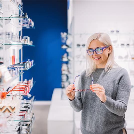 A woman with blonde hair and glasses smiles while holding eyeglasses in a store, surrounded by shelves of eyewear.