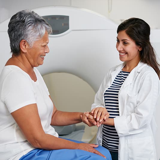A doctor in a white coat smiles and holds hands with an older woman sitting in front of a medical scanner, creating a supportive atmosphere.