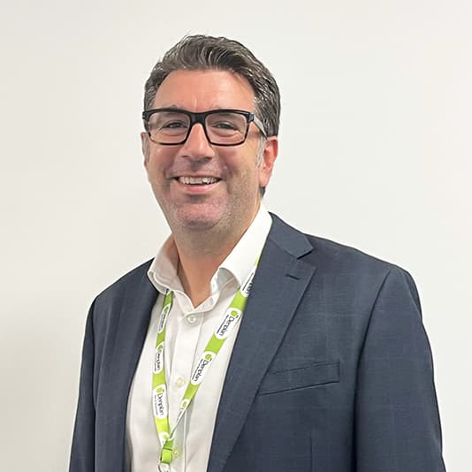 Smiling man in a suit with glasses and a lanyard stands against a plain white background.