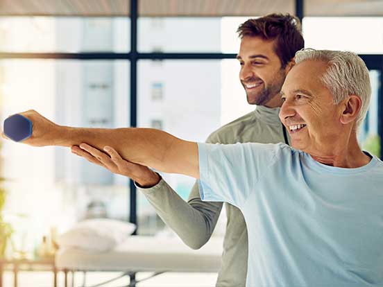 Older man smiling while lifting a dumbbell with assistance from a younger man in a bright, modern gym setting.