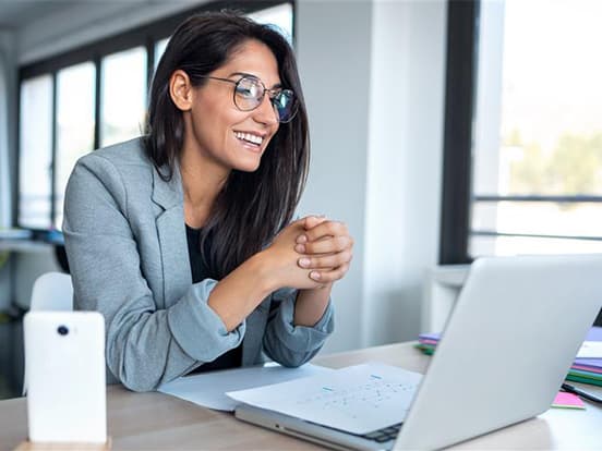Smiling woman in glasses and a blazer sits at a desk with a laptop, engaging in a video call in a bright office setting.